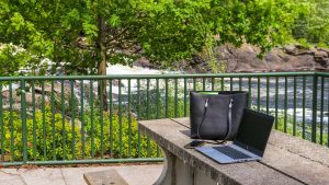 A laptop computer, a cell phone and a leather purse sitting on a concrete picnic table with a Muskoka waterfall and greenery in the background.
