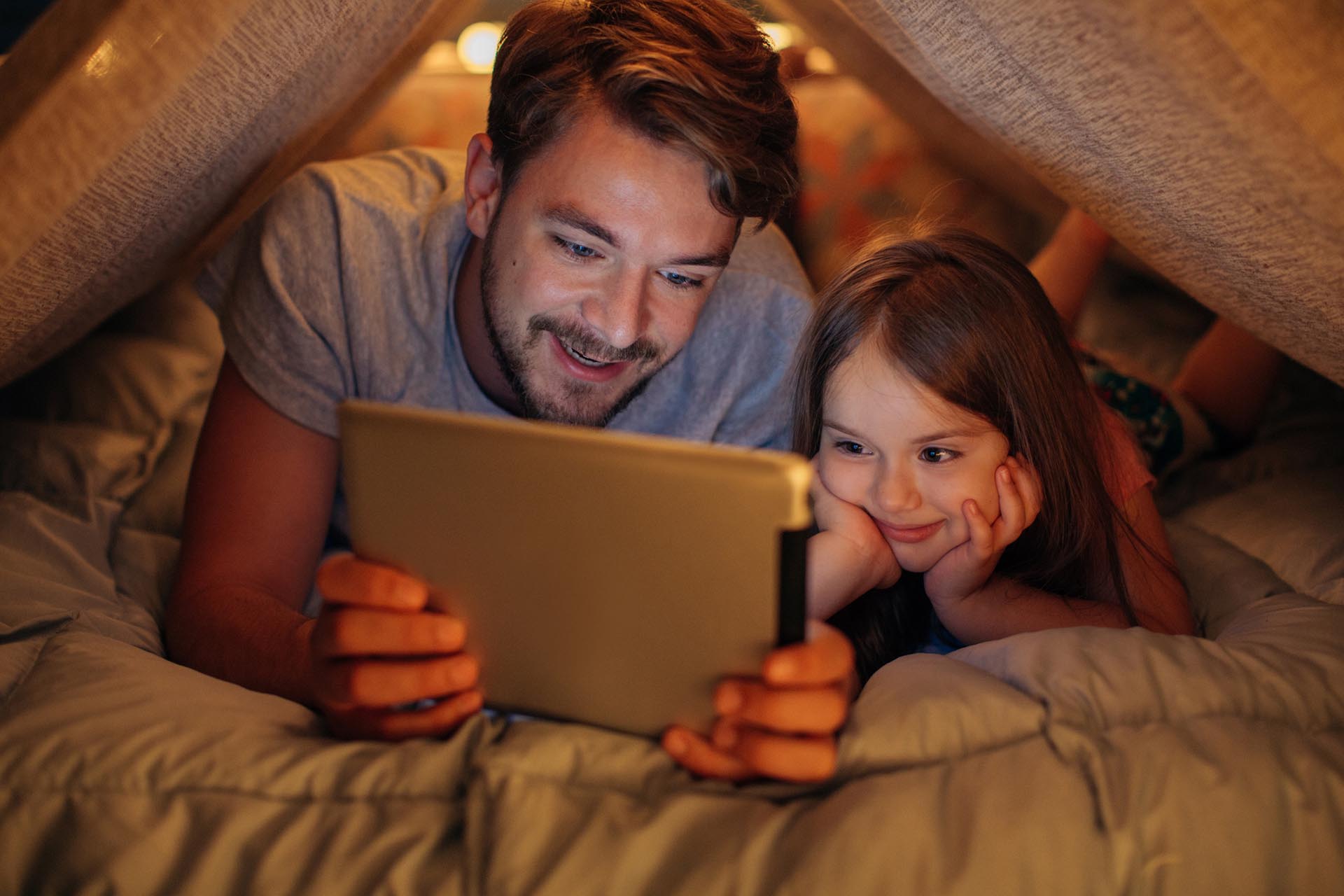 Father and daughter enjoying at home. Father and daughter in a blanket fort watching a digital tablet together.