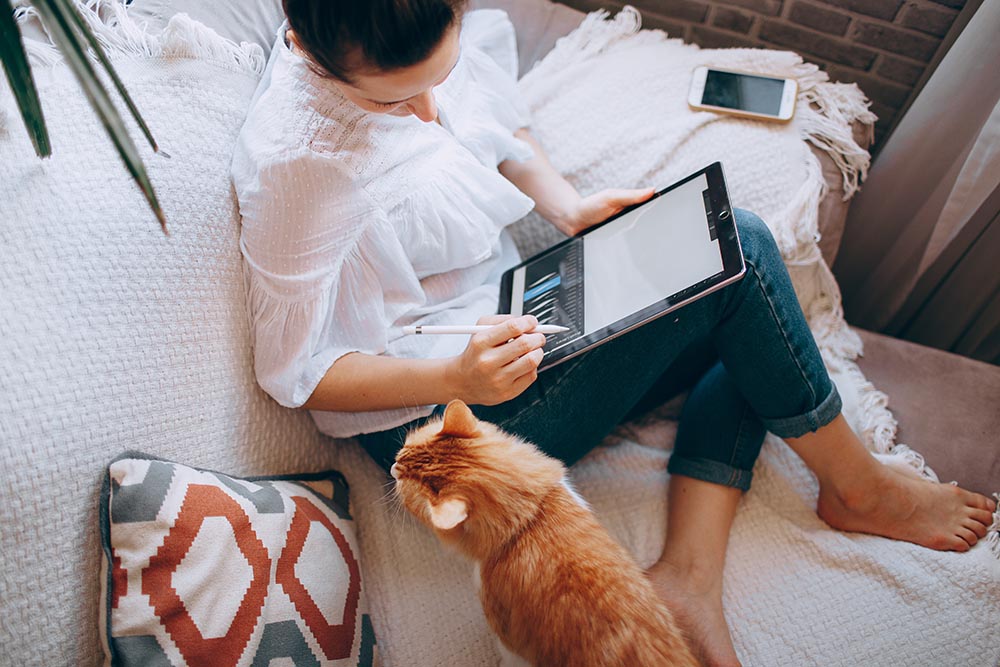 A woman sits on her couch drawing on a tablet with her orange cat 