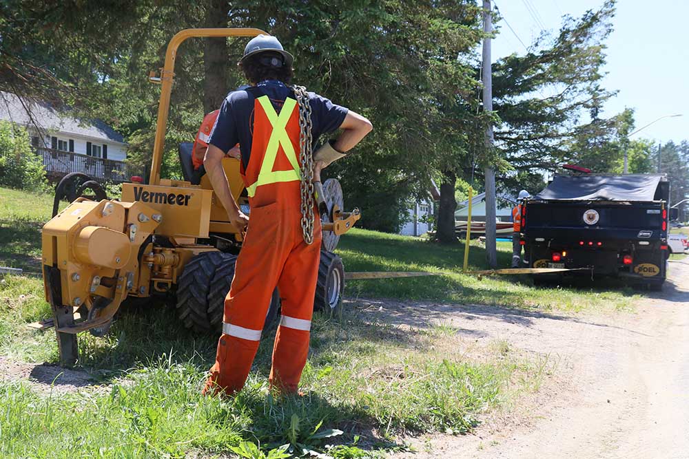 A Lakeland Networks technician wearing safety gear stands with his back to the camera, while another technician operates digging equipment to install fibre internet cables.