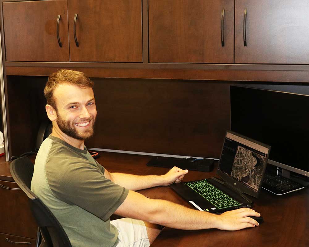 Lakeland Networks employee K.L. sits at a desk chair in front of a laptop with map software open on it. He is smiling at the camera.