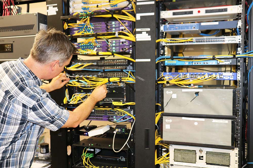 A Lakeland Networks employee, wearing a plaid shirt, adjusts yellow ethernet cables.