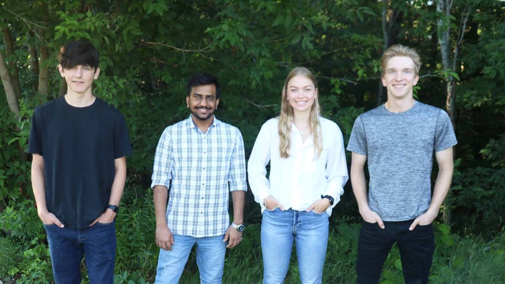 A group of Lakeland Networks summer students, three men and a woman, stand together, smiling, in front of a backdrop of trees. 
