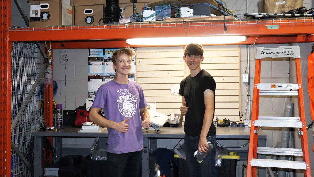 Two summer students, both men, smile and give the thumbs up in front of a work bench with tools and a step ladder. 