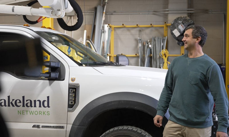 A Lakeland employee in a green shirt stands near a Lakeland Networks pickup truck.