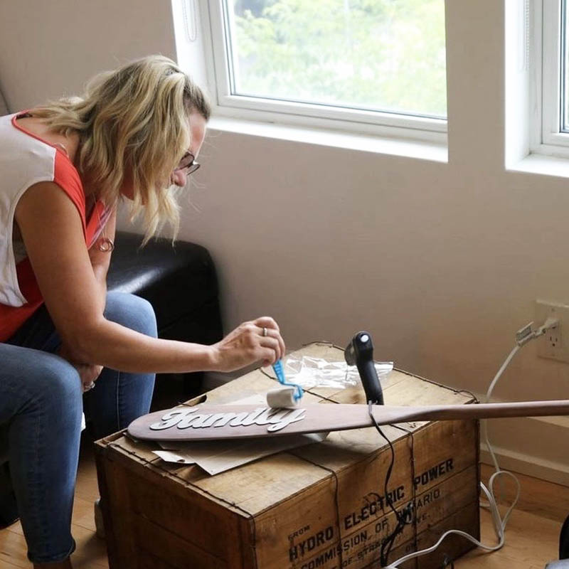 A blond woman rolls paint onto a wood cutout of the word "family" which is glued onto a wooden paddle. 