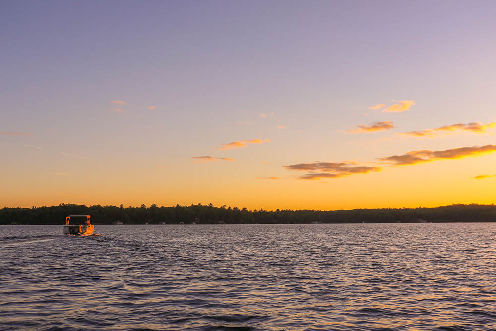A pontoon boat with a canopy cruises on a lake at sunset with the shoreline in the distance