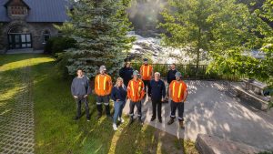 A group of smiling Lakeland Networks staff stand in front of falls in downtown Bracebridge. 4 are wearing orange safety vests and hard hats, and 4 are wearing blue sweatshirts.