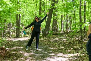 A young woman with dark hair and sunglasses, wearing black athletic clothing and standing on a trail in a forest, smiles and poses with her hands in the air