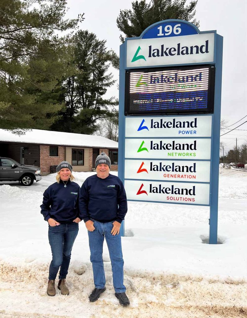 A woman and a man stand in front of the Lakeland Networks sign wearing Coldest Night of the Year toques.