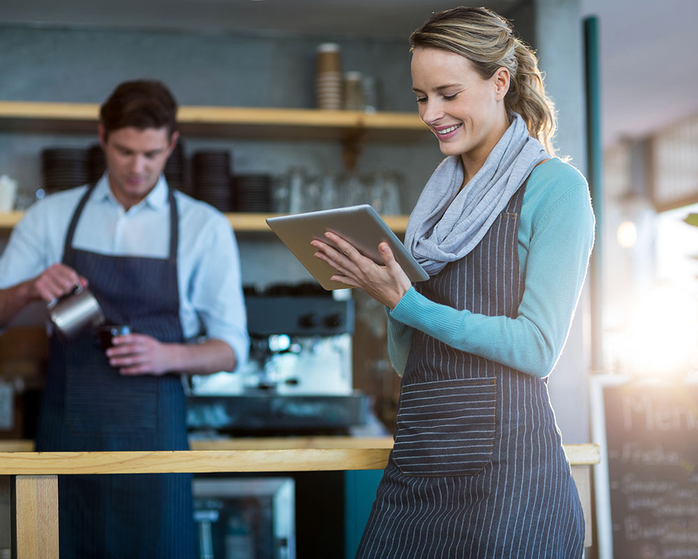 A blonde woman in a blue apron smiles and looks at a tablet, while a man in an apron pours a coffee in the background.