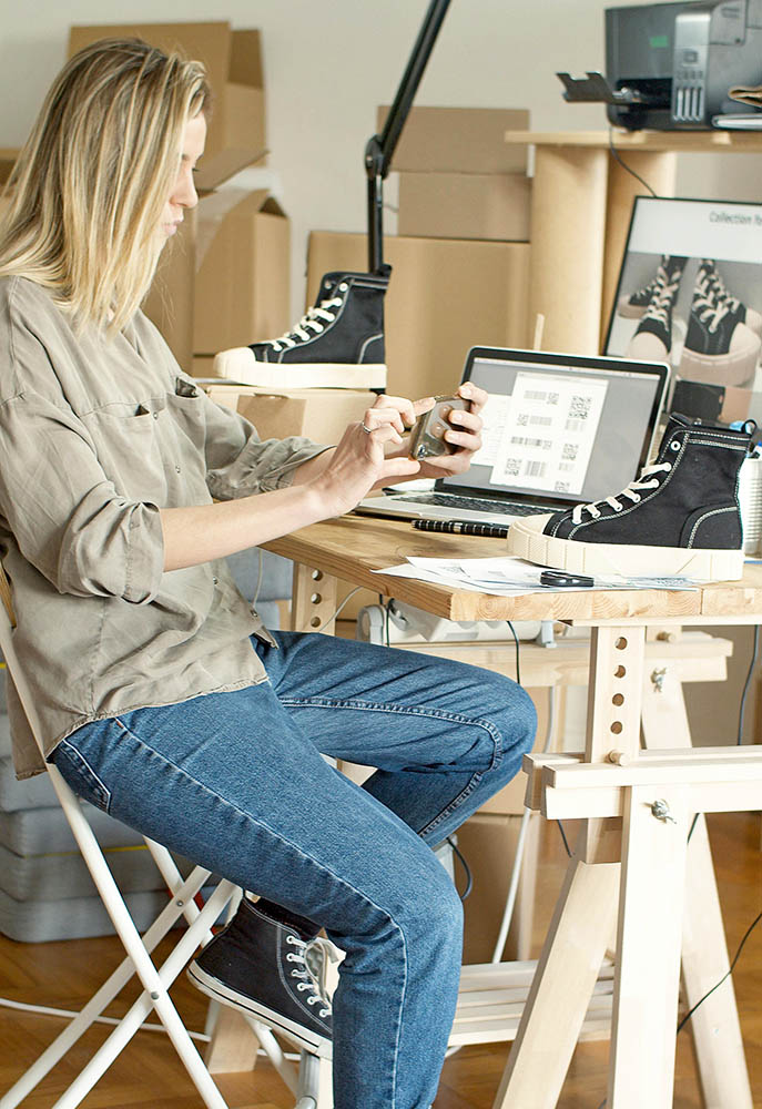 A blonde woman takes a photo of a shoe with a cell phone camera, with a desk and laptop in the background.